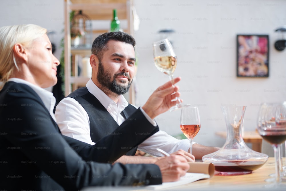 Young successful cavist looking at white wine held by his colleague during examination of its characteristics