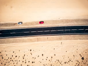 Aerial view of black straight asphalt road with sand and desert on both sides around - two car parked on the side - concept of travel and wanderlust for exotic destinations