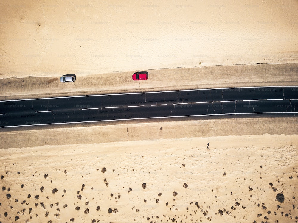両側に砂と砂漠がある黒いまっすぐなアスファルト道路の空撮 – 2台の車が側面に駐車 – エキゾチックな目的地への旅行と放浪癖のコンセプト