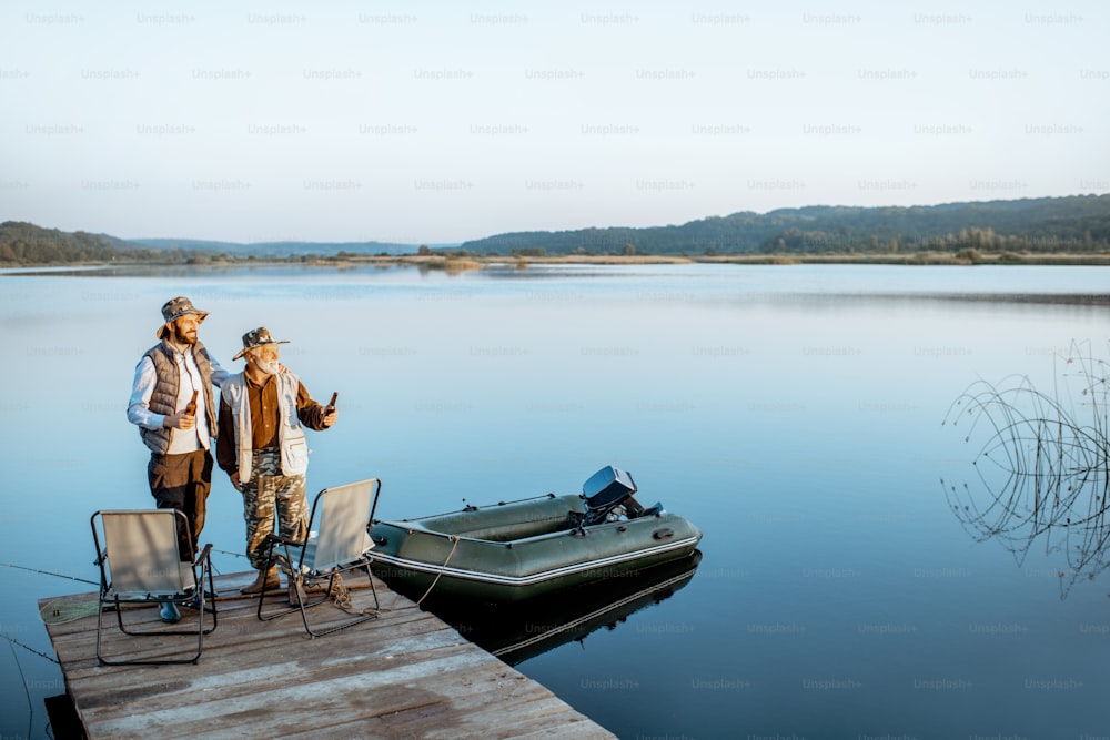 Grandfather with adult son standing together on the wooden pier, enjoying the sunrise while fishing on the lake early in the morning