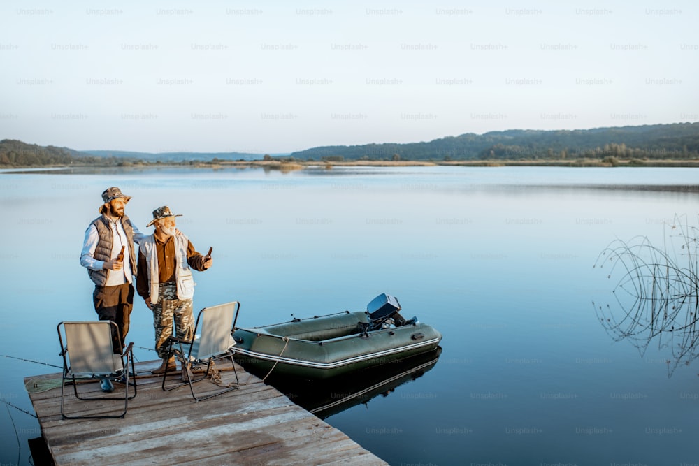 Avô com filho adulto em pé juntos no cais de madeira, apreciando o nascer do sol enquanto pescam no lago no início da manhã