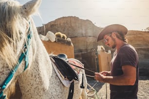 Young cute caucasian man in farm life cowboy style prepare her best friend white horse to go riding together enjoying the outdoor leisure activity and natural life with animals
