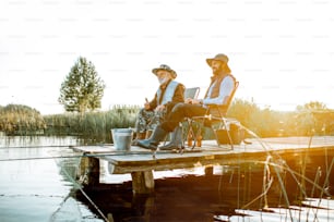 Grandfather with adult son fishing together on the wooden pier during the morning light. View from the side of the lake