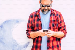 Coloured hipster people adult man with beard usgin modern cellular smart phone - standing on a white wall background - alternative job and work office in outdoor
