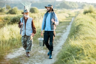 Grandfather with adult son walking with fishing gear on the footpath near the lake during the morning light