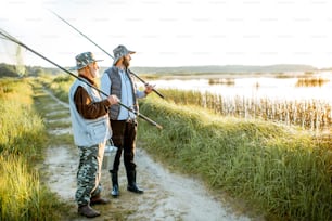 Grandfather with adult son standing together with fishing gear, enjoying landscape view on the lake during the morning light