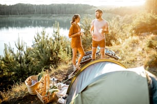Young couple standing at the campsite, talking and enjoying hot drinks while traveling in the mountains near the lake on the sunset