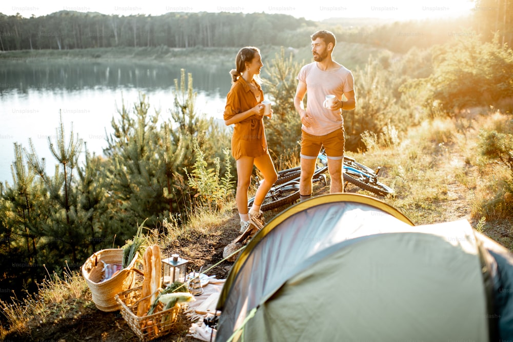 Jeune couple debout au camping, discutant et dégustant des boissons chaudes tout en voyageant dans les montagnes près du lac au coucher du soleil