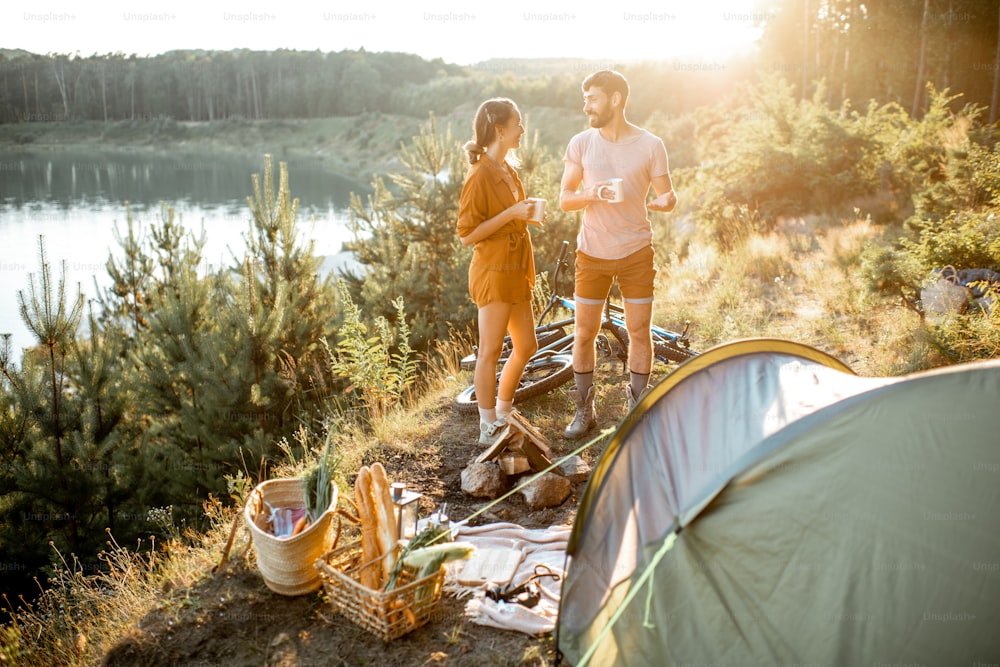 Jeune couple debout au camping, discutant et dégustant des boissons chaudes tout en voyageant dans les montagnes près du lac au coucher du soleil