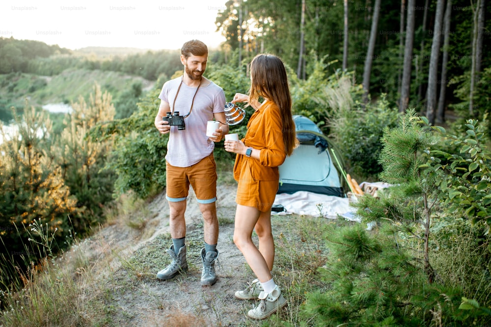 Jeune couple debout avec des boissons chaudes au camping dans la forêt, profitant de leur voyage d’été au coucher du soleil