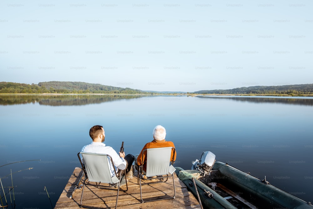 Grandfather with adult son enjoying beer, sitting together on the pier while fishing on the lake early in the morning. Wide landscape view