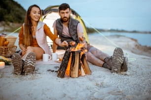 Coppia giovane e allegra che cucina salsicce al camino, facendo un picnic al campeggio sulla spiaggia la sera