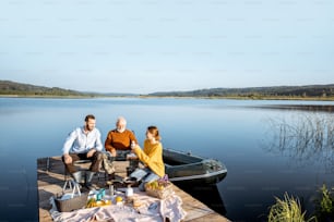 Mann und Frau mit älterem Großvater beim Picknick mit Gemüse und frisch gefangenem Fisch auf dem See am Morgen. Weite Landschaftsansicht