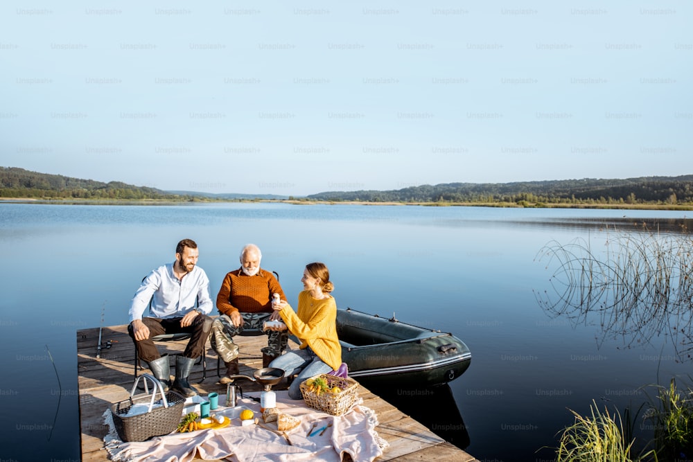 Man and woman with senior grandfather having a picnic with vegetables and fresh caught fish on the lake in the morning. Wide landscape view