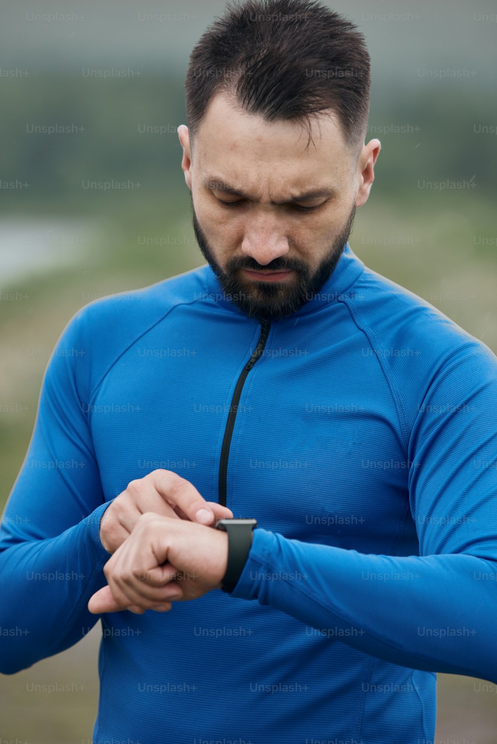 Portrait of mid age bearded sportsman exercising outdoor in summer, on gloomy day, standing at the road with scenic view, checking his smart watch, wearing blue shirt
