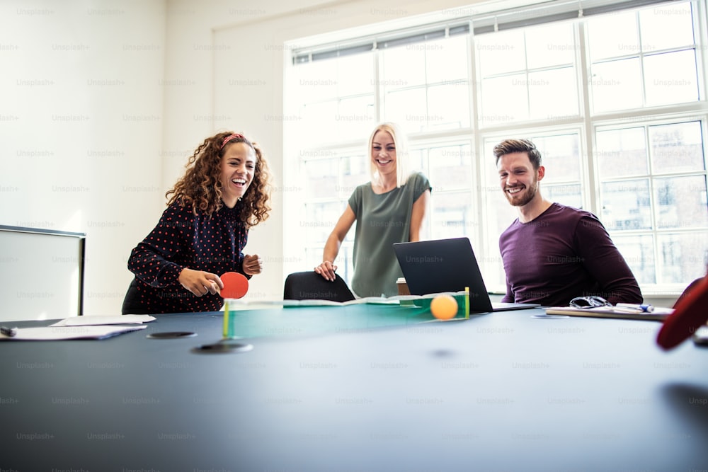 Laughing group of young designers playing table tennis on a boardroom table during a break from an office meeting