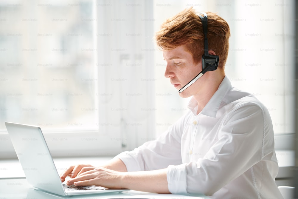 Jeune opérateur de centre d’appels communiquant avec des clients en ligne assis devant un ordinateur portable au bureau
