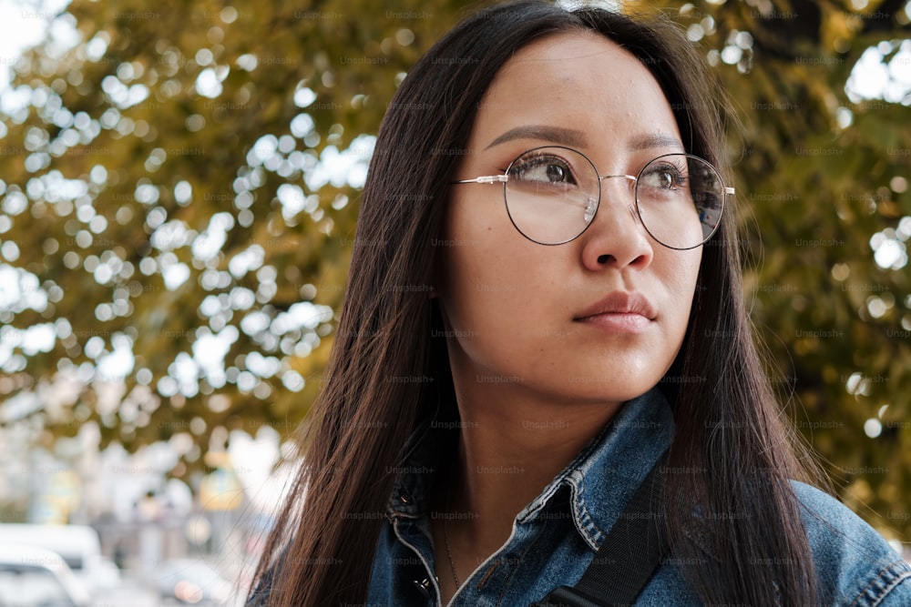 Close up portrait of young korean girl wearing round glasses and blue old school jeans, outdoors in summer, looking away from camera