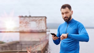 Portrait of mid age bearded sportsman checking his smart watch with smart phone, synchronizing, exercising outdoor in summer, on gloomy day with scenic view, wearing blue shirt