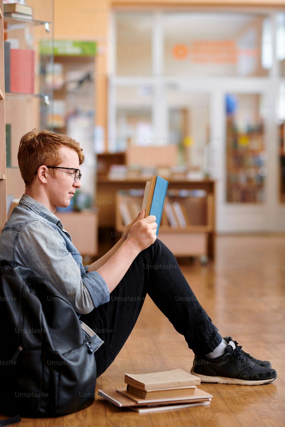 캐주얼 복장을 입은 젊은 영리한 남자가 수업이 끝난 후 도서관 바닥에 앉아 책을 읽고 있습니다.
