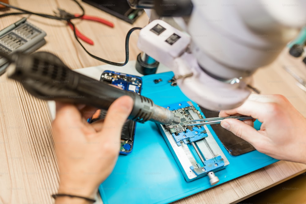 Herramienta de mano eléctrica y pinzas en manos del reparador que usa el microscopio durante el trabajo con pequeños detalles de teléfono inteligente desmontado
