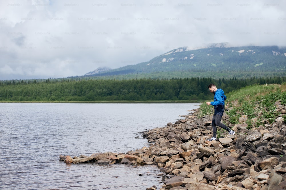 夏の屋外の岩だらけの湖の土手でジョギングするスポーツマン、湖と山の美しい景色を望む暗い日に、青いレインコートを着て、全長
