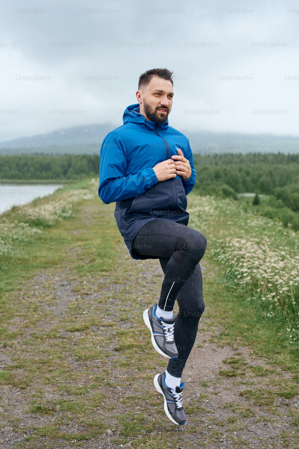 Mid age sportsman exercising outdoor in summer, on gloomy day, standing at the road with scenic view, doing warm up stretching, wearing blue jacket, he has a beard