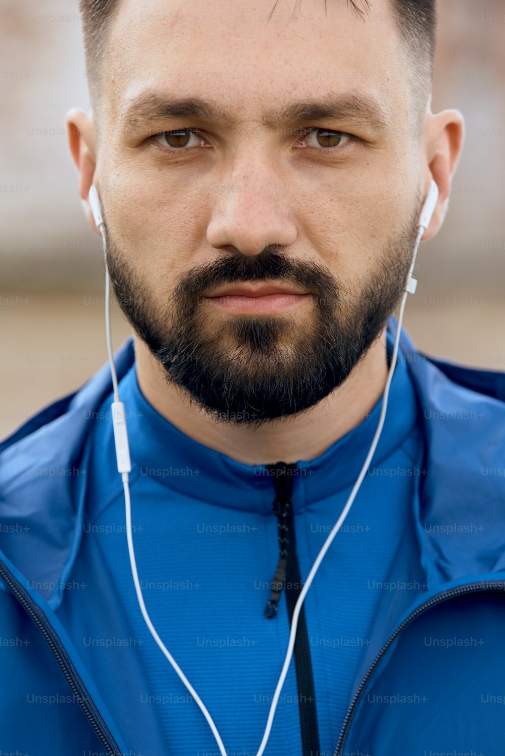 Portrait of mid age bearded sportsman with white earphones outdoor in summer, on sunny day with scenic view, wearing blue shirt, looking at the camera