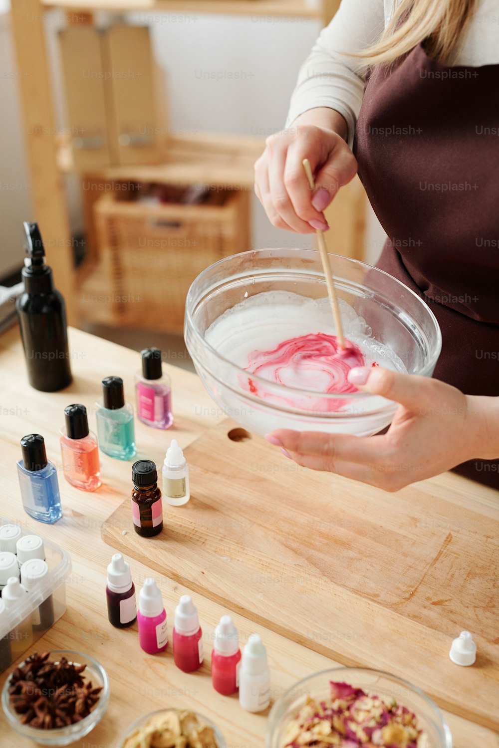 Hands of craftswoman mixing liquid soap mass with crimson color in glassware with wooden stick over table