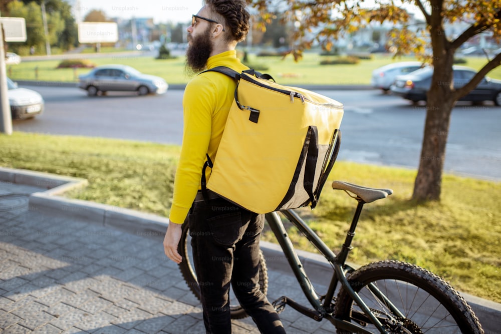 Young courier delivering food in yellow thermo bag, walking with bicycle in the city. Delivery service concept