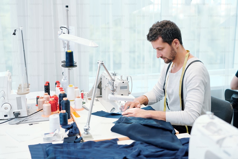 Young man concentrating on sewing clothing item on machine while working over order of client