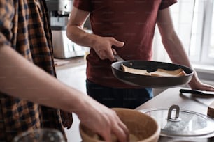 Close up of two men cooking breakfast together in the kitchen, one man is holding a pan, another man is preparing toasts