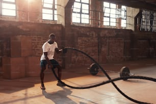 Fitness workout. Sport man doing battle rope exercise at gym. Black male athlete exercising, doing functional fitness training with heavy ropes indoors