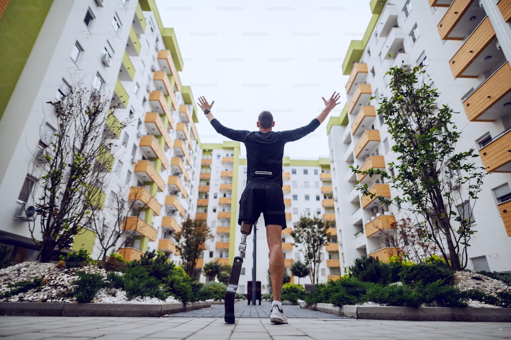 Vue arrière d’un beau sportif avec une jambe artificielle debout avec les mains en l’air à l’extérieur entouré de bâtiments.