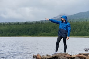 Sportsman standing on rocky lake bank, pointing forward outdoor in summer, on gloomy day with scenic view, looking at the lake and mountains, wearing blue rain coat, full length