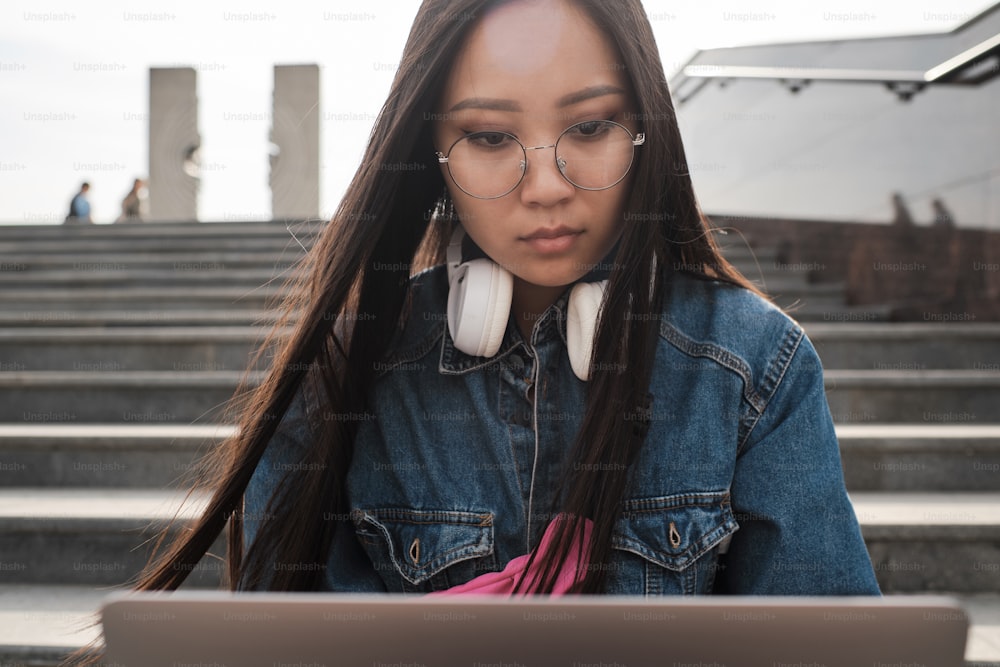 Chica asiática con gafas y auriculares mira la pantalla de la computadora portátil con firmeza.