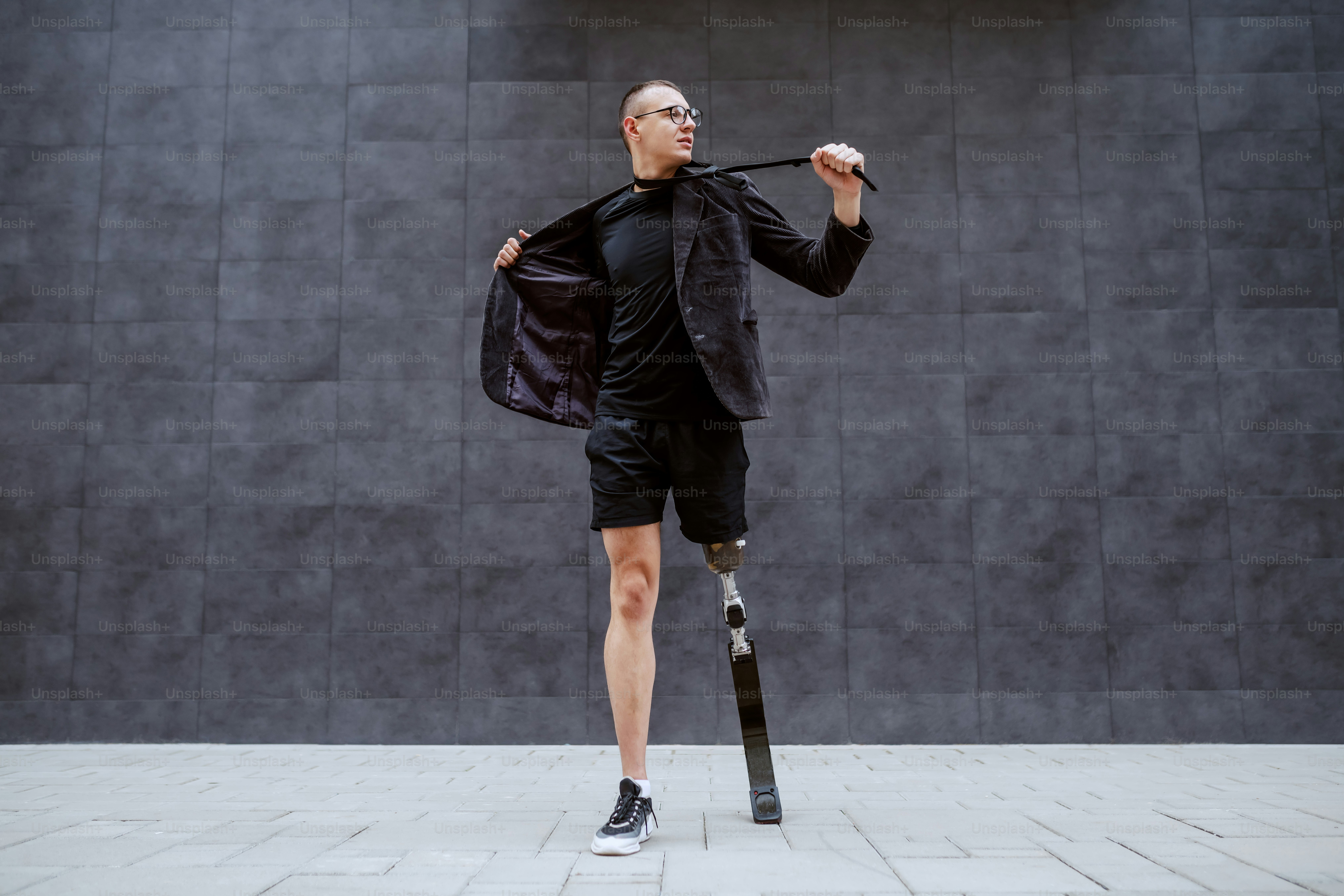 De corpo inteiro de belo empresário caucasiano com perna artificial se cansando do trabalho e rasgando seu terno enquanto estava ao ar livre em frente à parede cinza. É hora do esporte.