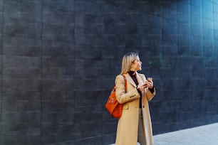 Affascinante donna bionda sorridente caucasica in cappotto e con borsa tenendo smart phone mentre si trova all'aperto. Sullo sfondo c'è un muro scuro.
