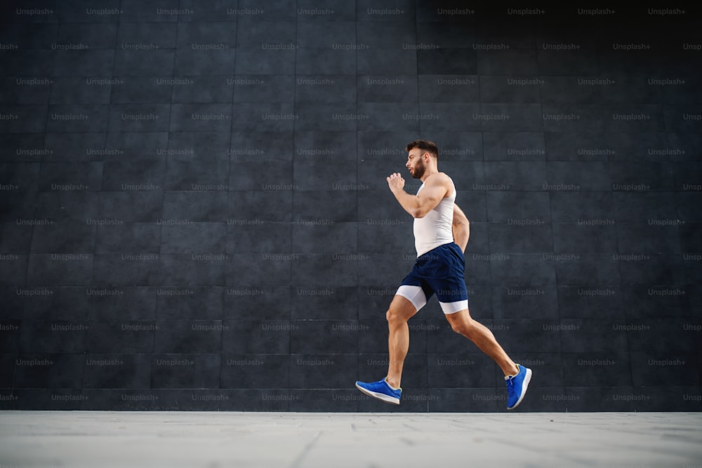Side view of handsome caucasian muscular fit man running fast outdoors. In background is gray wall.