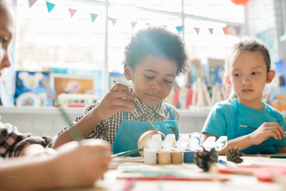 Serious schoolboy and his classmate sitting by desk and painting Christmas decorations with gouache at lesson