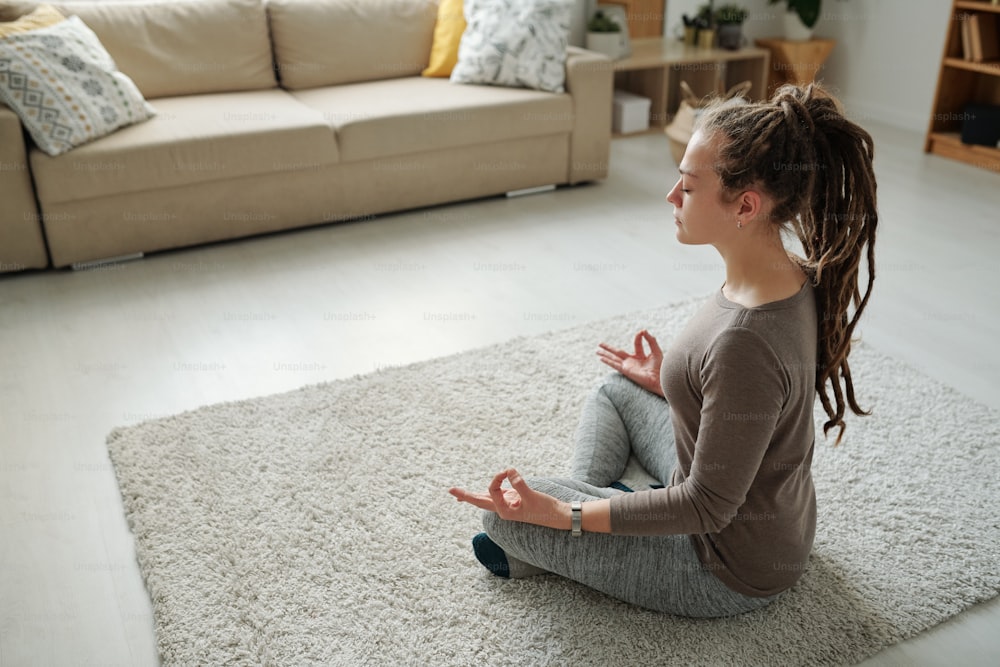 Fit girl in activewear listening sitting in pose of lotus on carpet while meditating during yoga practice