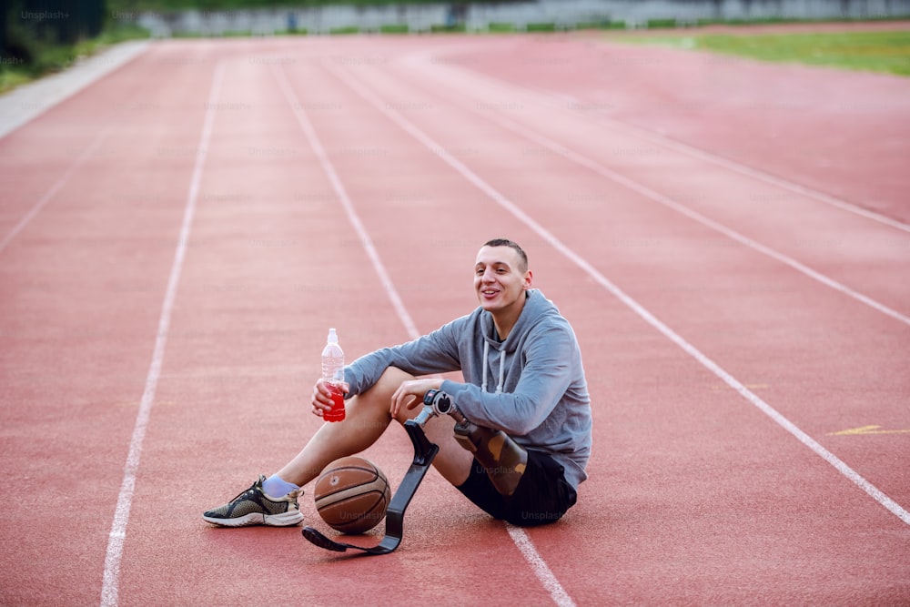 Uomo handicappato sportivo caucasico altamente motivato in abbigliamento sportivo seduto in pista e in possesso di rinfresco. Tra le gambe c'è la palla da basket.