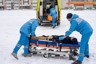 Dois paramédicos em trajes de trabalho azuis consertando homem inconsciente com cintos na maca para empurrá-lo então para o carro de ambulância