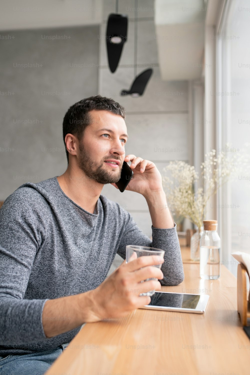 Hübscher junger Mann im lässigen grauen Pullover beim Smartphone, während er durch das Fenster in einem gemütlichen Café schaut