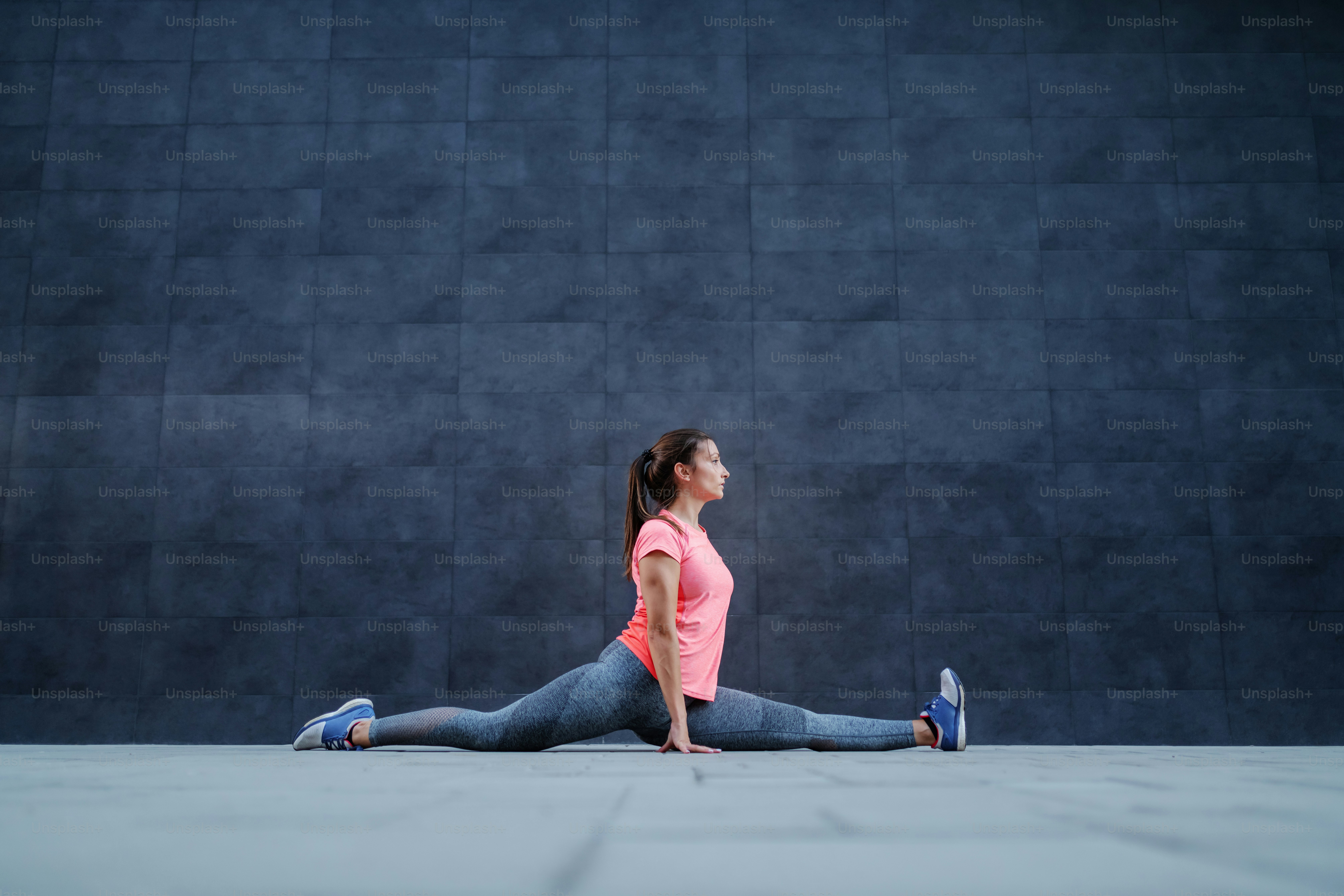 Ajuste atractivo deportista caucásico flexible en ropa deportiva haciendo divisiones al aire libre. Al fondo hay una pared oscura.
