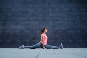 Fit attractive flexible caucasian sportswoman in sportswear doing splits outdoors. In background is dark wall.