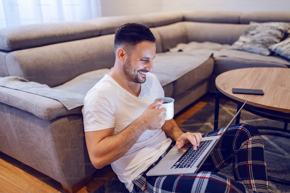 Belo homem caucasiano sem barba sorridente sentado no chão da sala de estar, navegando na internet e bebendo café pela manhã.