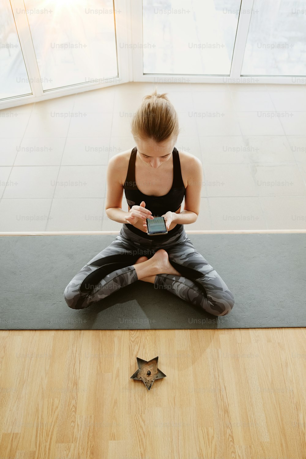 Full body shot of young skinny woman meditating while sitting on mat in front of big bright window, burning scented sticks. She is wearing tank and dark pants