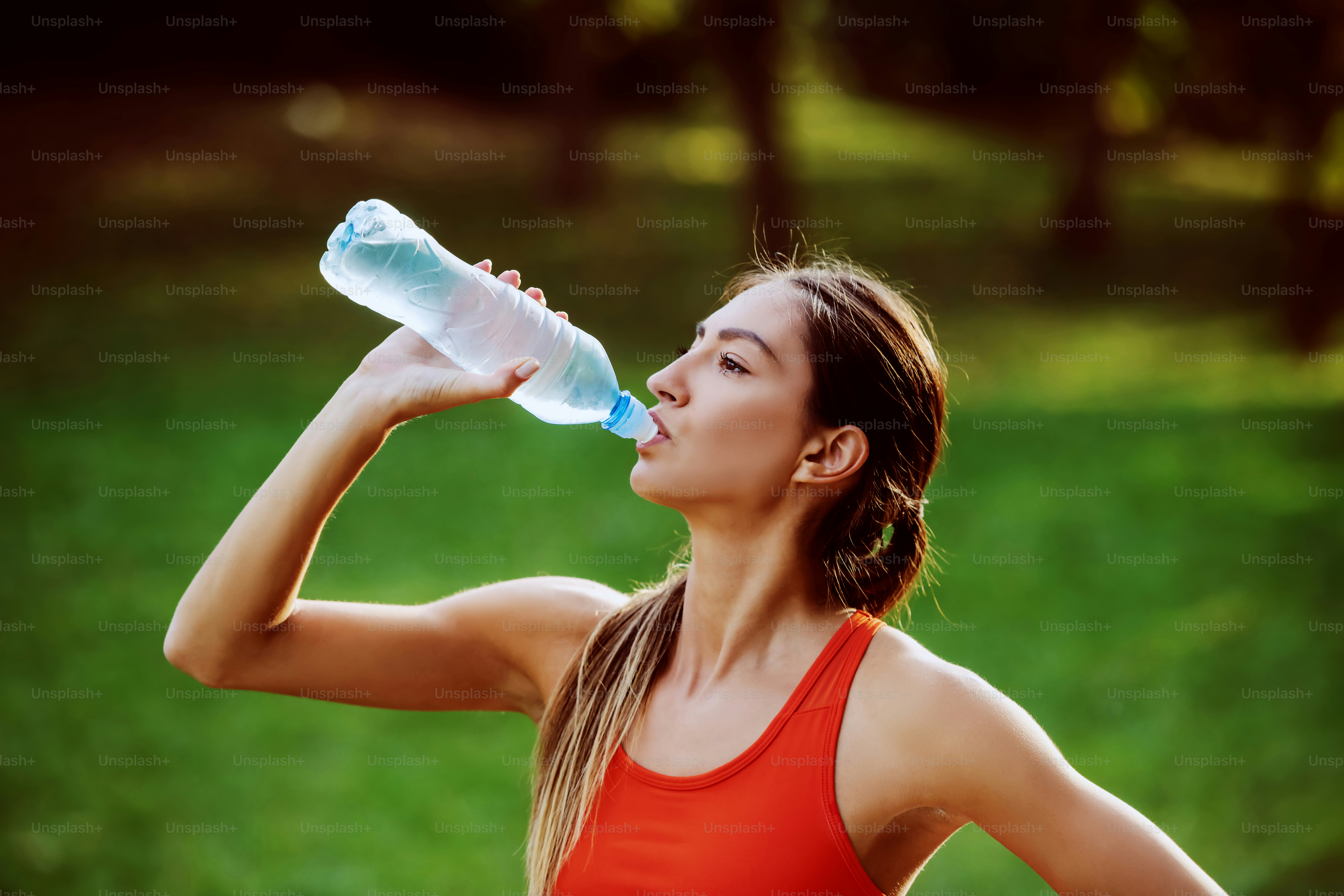 Attractive thirsty caucasian fit brunette in sportswear and with ponytail standing in nature and drinking water. Sunny day in nature.