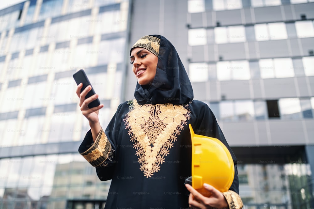 Vista a basso angolo di attraente donna musulmana sorridente in piedi di fronte all'edificio aziendale, usando lo smartphone e tenendo il casco sotto l'ascella. Anche le donne possono essere grandi architetti.